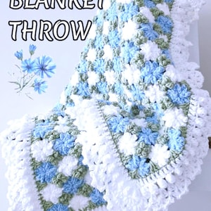 Lavishly beautiful crochet floral blanket made of cornflower motifs is hanging from a display. The blanket resembles a field of white and blue cornflowers framed with a beautiful lace edging.  The lettering says "Cornflowers Blanket Throw", "Crochet Pattern By JTEasyCrochet" meaning that JTEasyCrochet has the best crochet patterns for flower baby blankets, throws and afghans.