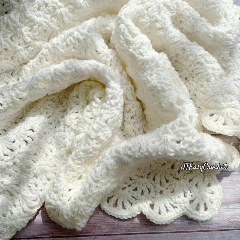 Cream-colored, handmade crochet blanket with a scalloped edge. The blanket features a detailed stitch pattern and is draped over a wooden surface. The text "JTEasyCrochet" is visible in the lower right corner meaning that JTEasyCrochet has the best crochet patterns, beautiful and easy.