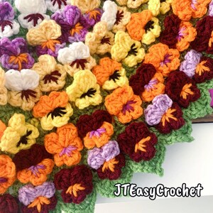 May include: A close-up of a crocheted blanket featuring colorful pansies. The flowers are in shades of white, yellow, orange, purple, and burgundy, with green leaves. The text "JTEasyCrochet" is visible in the bottom right corner.