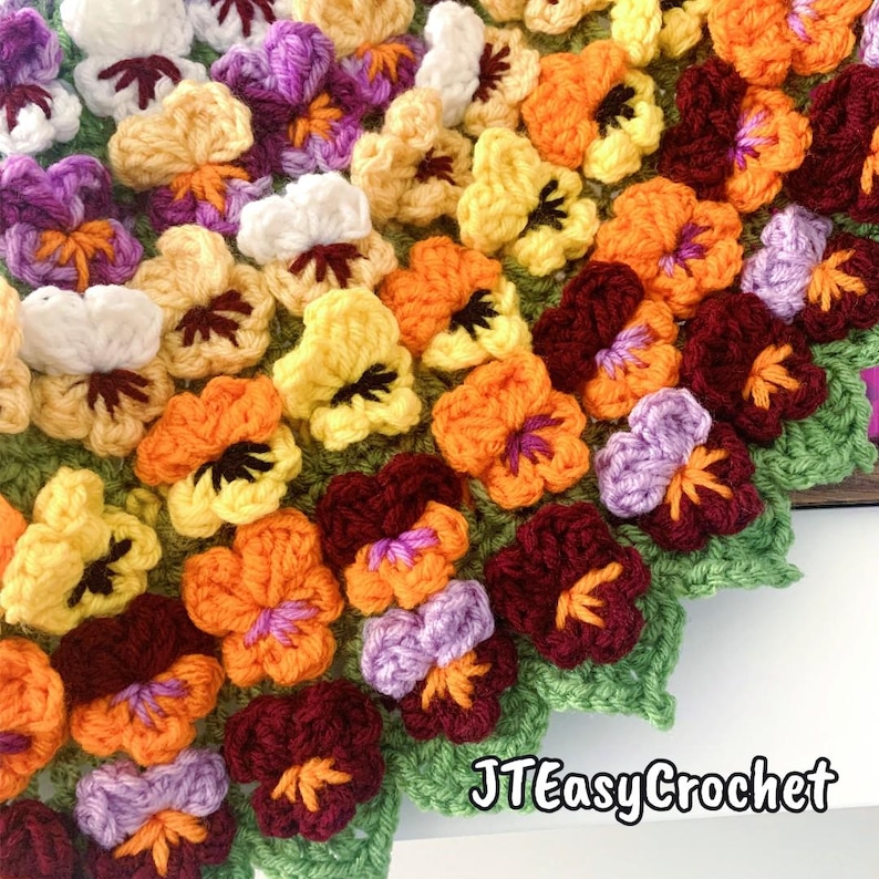 May include: A close-up of a crocheted blanket featuring colorful pansies. The flowers are in shades of white, yellow, orange, purple, and burgundy, with green leaves. The text "JTEasyCrochet" is visible in the bottom right corner.
