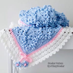 May include: A handmade, crocheted baby blanket in shades of light blue, white, and pink. The blanket features a floral pattern, a white scalloped edge, and a pink border. A pink bow and two small flower accents are attached.