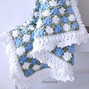 Beautiful blue and white crochet floral blanket made of cornflower motifs is hanging from a display against a light gray surface. The blanket resembles a field of white and blue cornflowers framed with a beautiful lace edging.  The lettering says "JTEasyCrochet" meaning that JTEasyCrochet has the best crochet patterns for flower blankets.