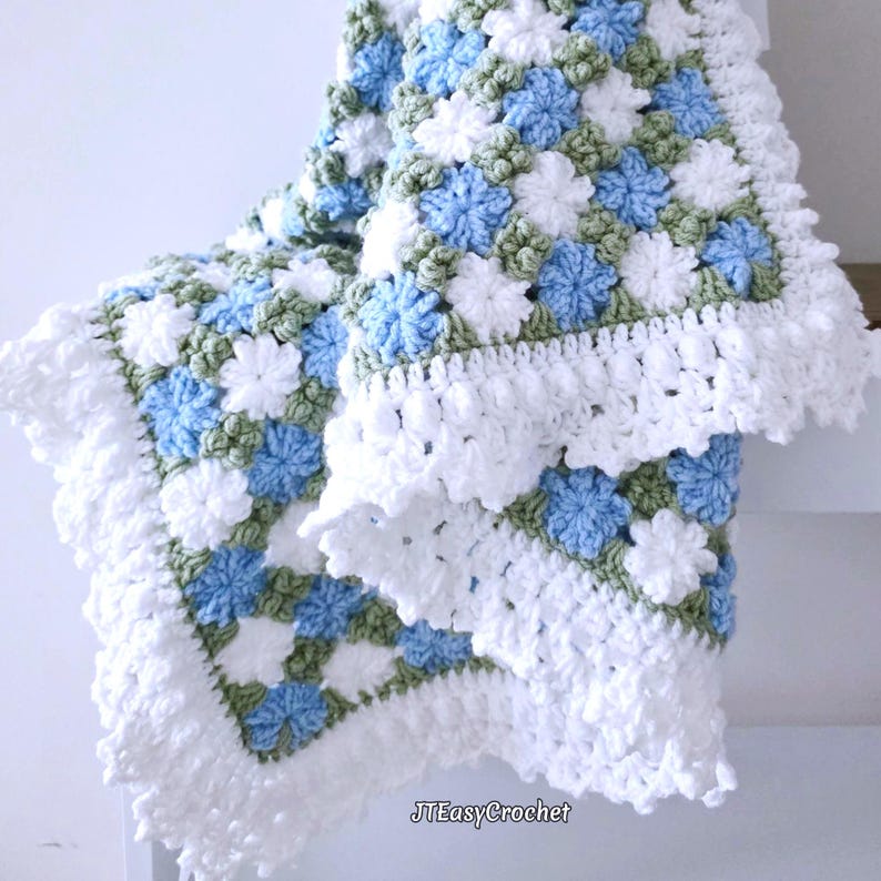 Beautiful blue and white crochet floral blanket made of cornflower motifs is hanging from a display against a light gray surface. The blanket resembles a field of white and blue cornflowers framed with a beautiful lace edging.  The lettering says "JTEasyCrochet" meaning that JTEasyCrochet has the best crochet patterns for flower blankets.