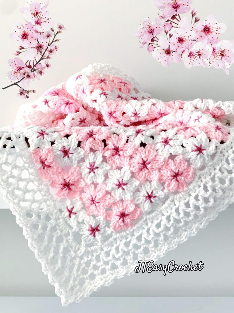 May include: A handmade crochet blanket in white, pink, and red. The blanket features a floral pattern with a white openwork border. The text "JTEasyCrochet" is visible in the lower right corner. Cherry blossom branches are in the background.