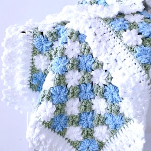 Lavishly beautiful crochet floral blanket made of cornflower motifs is hanging from a display. The blanket resembles a field of white and blue cornflowers framed with a beautiful lace edging.  The lettering says "Cornflowers Baby Blanket", "Crochet Pattern By JTEasyCrochet" meaning that JTEasyCrochet has the best crochet patterns for flower baby blankets, throws and afghans.