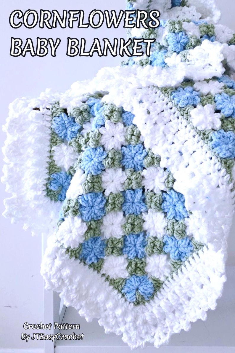 Lavishly beautiful crochet floral blanket made of cornflower motifs is hanging from a display. The blanket resembles a field of white and blue cornflowers framed with a beautiful lace edging.  The lettering says "Cornflowers Baby Blanket", "Crochet Pattern By JTEasyCrochet" meaning that JTEasyCrochet has the best crochet patterns for flower baby blankets, throws and afghans.