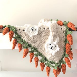 Puede incluir: Una manta de beb&eacute; de ganchillo con fondo beige y una aplicaci&oacute;n de conejito blanco. La manta est&aacute; adornada con hojas verdes y zanahorias naranjas.