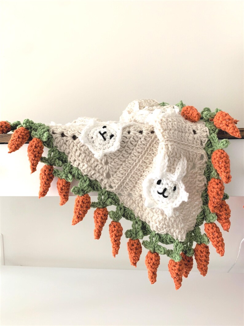 Puede incluir: Una manta de beb&eacute; de ganchillo con fondo beige y una aplicaci&oacute;n de conejito blanco. La manta est&aacute; adornada con hojas verdes y zanahorias naranjas.