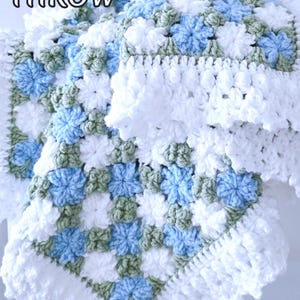 Lavishly beautiful crochet floral blanket made of cornflower motifs is hanging from a display. The blanket resembles a field of white and blue cornflowers framed with a beautiful lace edging.  The lettering says "Cornflowers Baby Blanket", "Crochet Pattern By JTEasyCrochet" meaning that JTEasyCrochet has the best crochet patterns for flower baby blankets, throws and afghans.