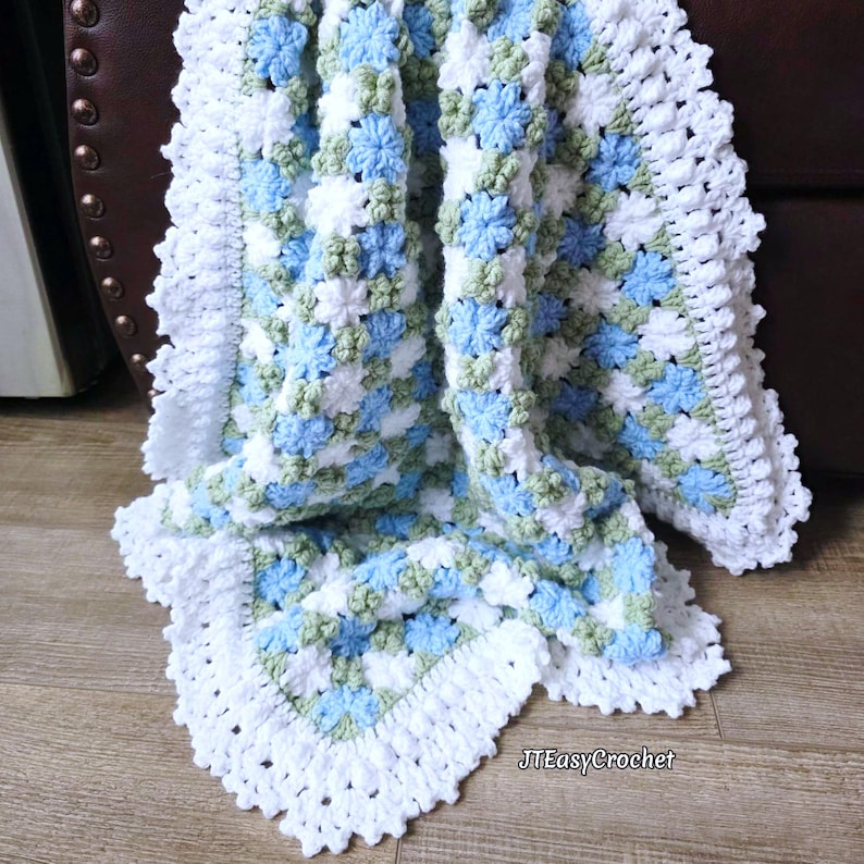 May include: A crocheted blanket with a granny square design. The blanket is made with white, light blue, and green yarn, and has a white, scalloped edge. The blanket is folded, displaying its texture and pattern.