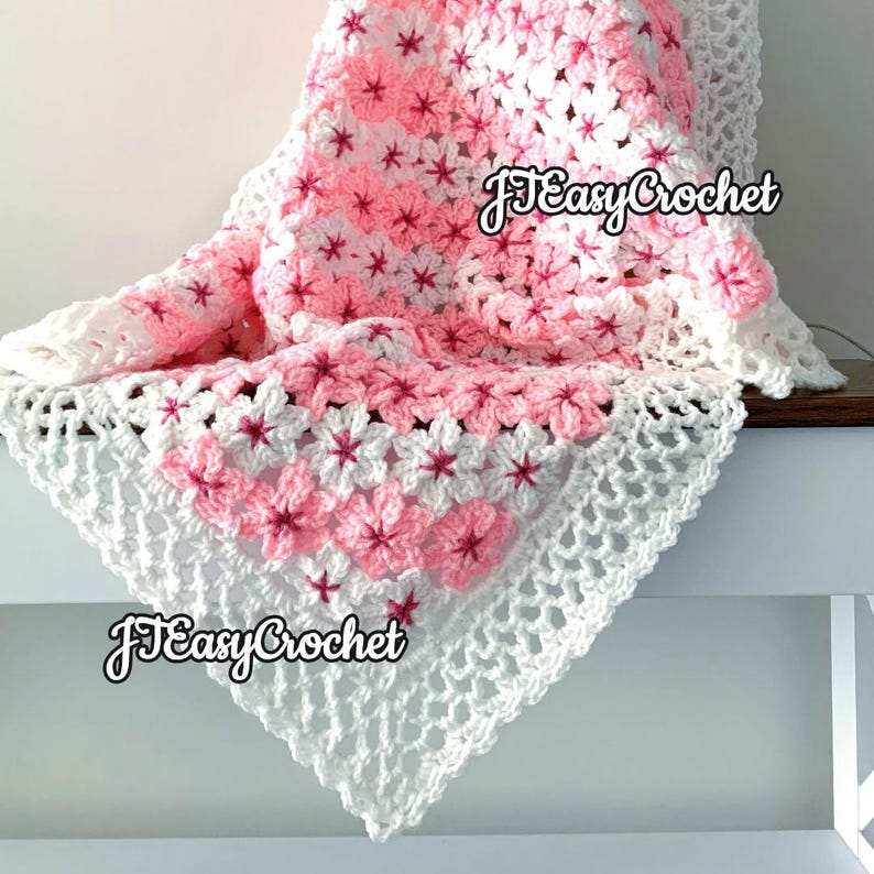 May include: A handmade crochet blanket featuring a floral pattern in shades of pink and white. The blanket has a delicate, openwork border and the text "JEasyCrochet" is visible. This cozy blanket is perfect for adding a touch of handmade charm to any space.