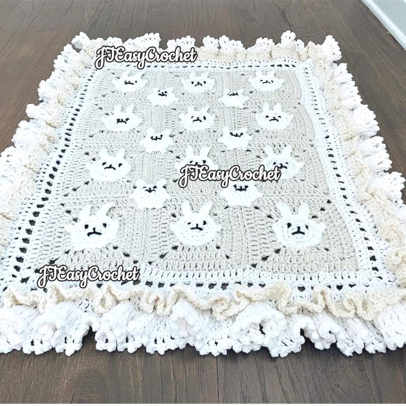 Op de afbeelding: Een gehaakt babydekentje met een witte rand en een beige middenstuk met een herhalend patroon van witte konijnengezichten. Het dekentje heeft een gegolfde rand en de tekst "JJ Easy Crochet" is zichtbaar op de afbeelding.