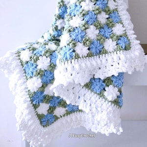 Lavishly beautiful crochet floral blanket made of cornflower motifs is hanging from a display. The blanket resembles a field of white and blue cornflowers framed with a beautiful lace edging.  The lettering says "JTEasyCrochet" meaning that JTEasyCrochet has the best crochet patterns for flower baby blankets, throws and afghans.