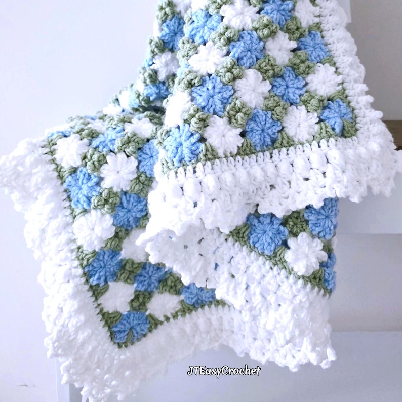 Lavishly beautiful crochet floral blanket made of cornflower motifs is hanging from a display. The blanket resembles a field of white and blue cornflowers framed with a beautiful lace edging.  The lettering says "JTEasyCrochet" meaning that JTEasyCrochet has the best crochet patterns for flower baby blankets, throws and afghans.