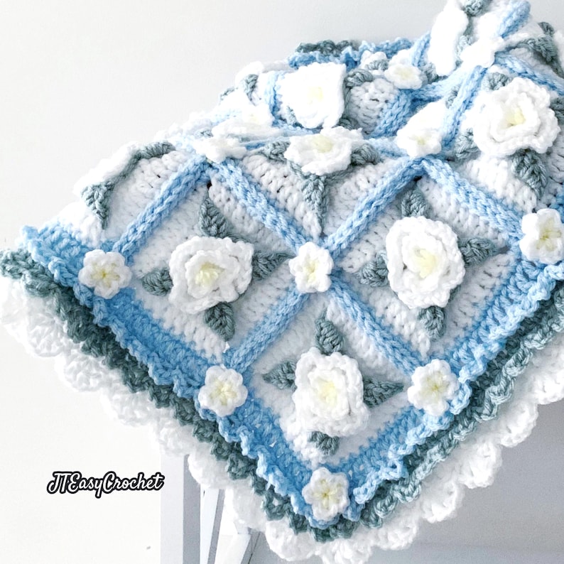 Beautiful crochet granny square blanket is hanging from a display on a light grey background. Each white granny square has a white textured gardenia flower in the center, framed with pale green leaves and light frosty blue border, so the whole blanket remind of a frozen gardenia blanket. There are also bunches of gardenia flowers in the image to highlight the likeness. The lettering logo says: " JTEasyCrochet"