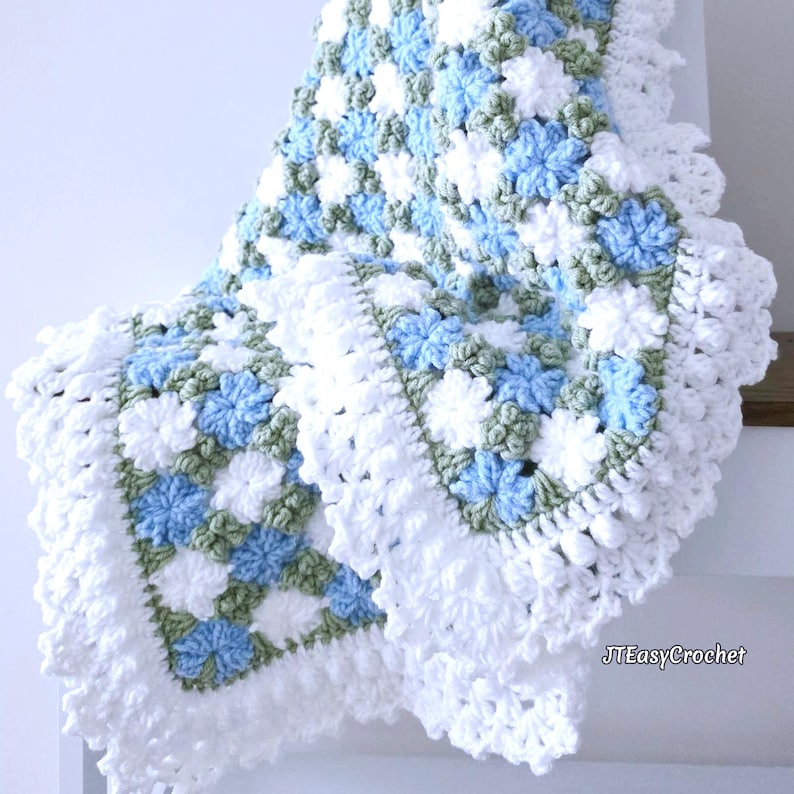 Beautiful blue and white crochet floral blanket made of cornflower motifs is hanging from a display. The blanket resembles a field of white and blue cornflowers framed with a beautiful lace edging.  The lettering says "JTEasyCrochet" meaning that JTEasyCrochet has the best crochet patterns for flower blankets.