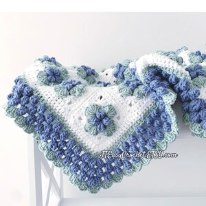 Beautiful white granny square blannket is laying on a white shelf on a white background. In the center of each square is blueberry with green leaves. The border is made of blueberries.