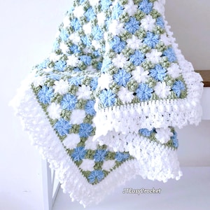 Lavishly beautiful crochet floral blanket made of cornflower motifs is hanging from a display. The blanket resembles a field of white and blue cornflowers framed with a beautiful lace edging.  The lettering says "JTEasyCrochet" meaning that JTEasyCrochet has the best crochet patterns for flower blankets.