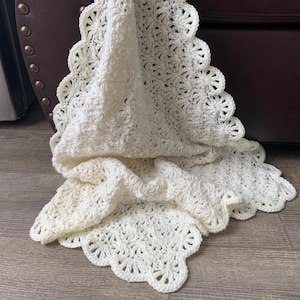 May include: Cream-colored, handmade crochet blanket with a scalloped edge. The blanket features a detailed floral pattern throughout and is draped over a dark brown surface. The soft texture and intricate design suggest a cozy, handcrafted item.