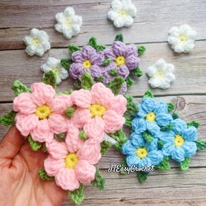 Op de afbeelding: Drie gebreide bloemclusters in roze, paars en blauw. Elke cluster bestaat uit meerdere individuele bloemen met gele centra. De bloemen zijn in een driehoekige vorm gerangschikt en hebben groene bladeren.