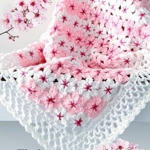 Pode incluir: Um cobertor de crochê feito à mão com um padrão floral em tons de rosa e branco. O cobertor apresenta motivos em forma de flor e uma borda vazada. O texto "JTEasyCrochet" é visível no canto inferior esquerdo.