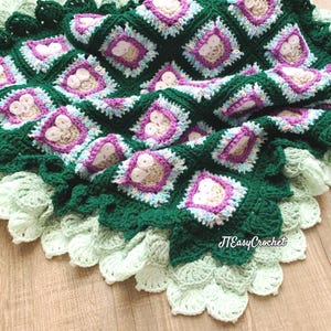 Puede incluir: Una manta de ganchillo hecha a mano en tonos verdes, blancos y morados. La manta presenta un patrón cuadrado repetido con diseños florales en el centro de cada cuadrado. El borde tiene un borde festoneado. El texto "JTEasyCrochet" es visible.