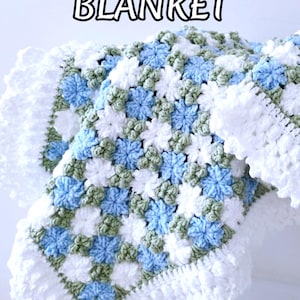 Lavishly beautiful crochet floral blanket made of cornflower motifs is hanging from a display. The blanket resembles a field of white and blue cornflowers framed with a beautiful lace edging.  The lettering says "Cornflower Blanket", "Crochet Pattern By JTEasyCrochet" meaning that JTEasyCrochet has the best crochet patterns for flower baby blankets, throws and afghans.