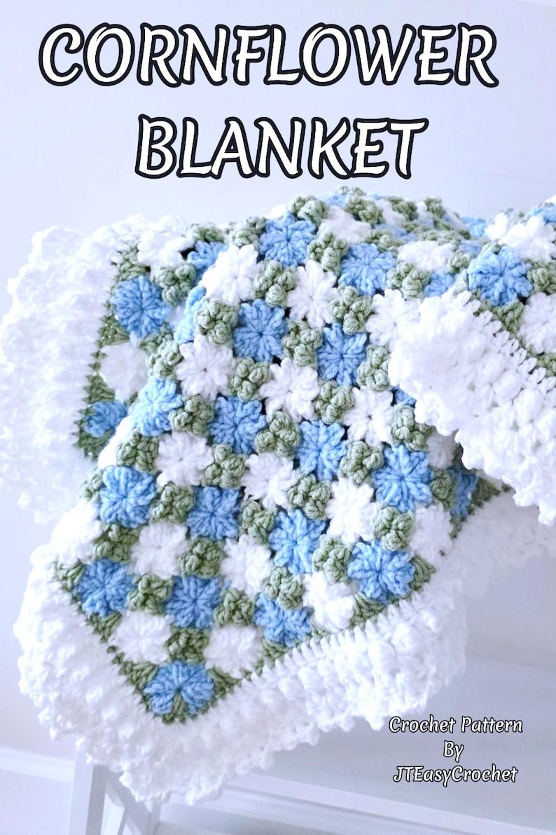 Lavishly beautiful crochet floral blanket made of cornflower motifs is hanging from a display. The blanket resembles a field of white and blue cornflowers framed with a beautiful lace edging.  The lettering says "Cornflower Blanket", "Crochet Pattern By JTEasyCrochet" meaning that JTEasyCrochet has the best crochet patterns for flower baby blankets, throws and afghans.