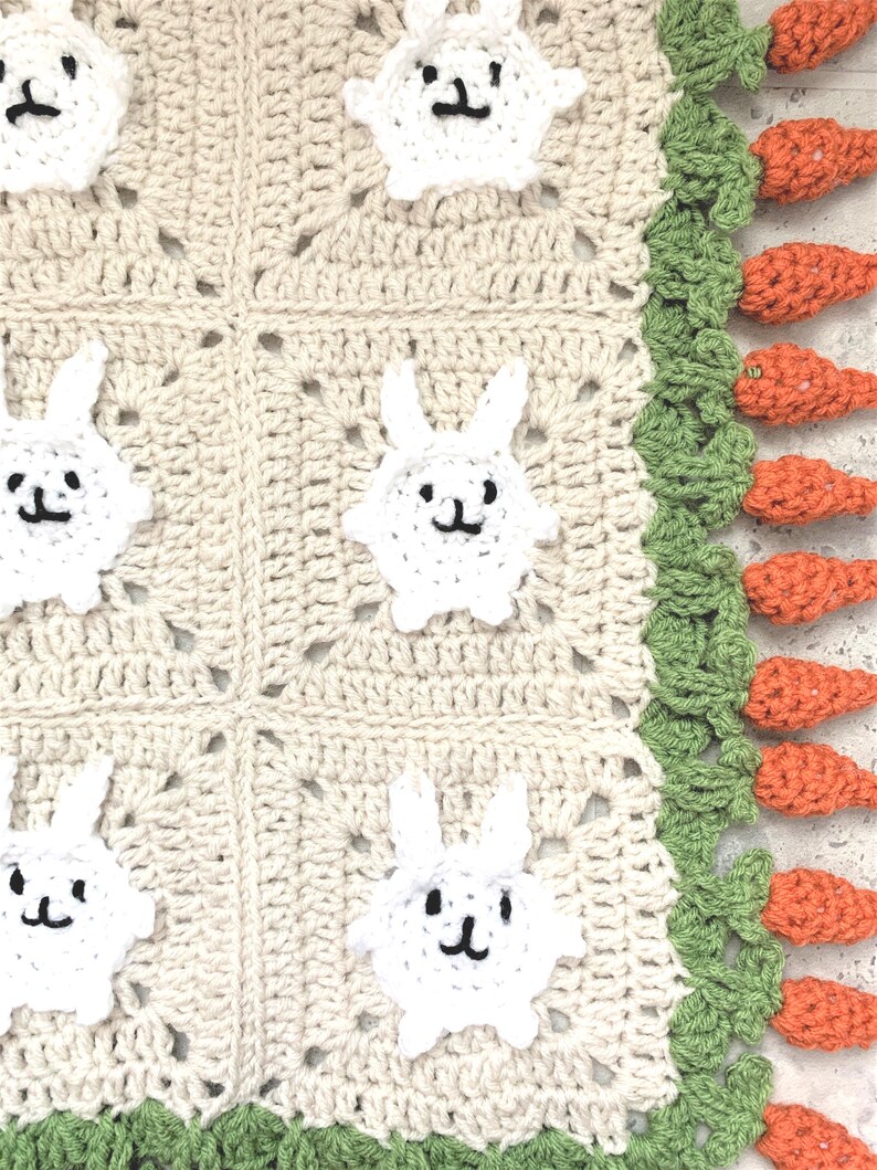 Puede incluir: Una manta de beb&eacute; de ganchillo con un dise&ntilde;o de conejito blanco y zanahoria. La manta es beige con ribetes verdes y naranjas.