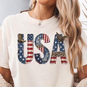 May include: An ivory short-sleeve t-shirt featuring the text USA in a distressed, multi-patterned design. The letters incorporate American flag motifs, denim textures, leopard print, and a small cowboy boot and eagle graphic accenting the letters.
