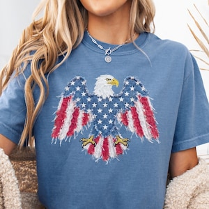 May include: Blue jean colored t-shirt featuring a distressed American flag eagle design. The eagle has a white head, yellow beak and talons, and wings with red and white stripes and blue stars. A casual, patriotic apparel item.