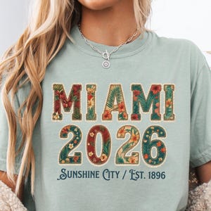 Puede incluir: Una camiseta de manga corta color verde salvia con el texto MIAMI 2026 en un patrón floral con un borde cosido color crema. Debajo del texto, las palabras Sunshine City / Est. 1896 están impresas en una fuente cursiva azul oscuro al frente.