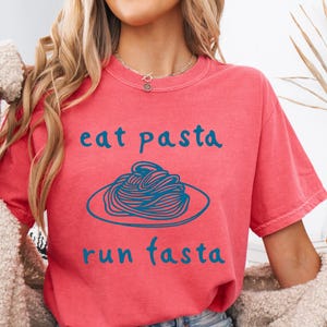 Peut inclure: T-shirt corail avec l'inscription bleue "eat pasta run fasta" et une illustration bleue de spaghettis. Le t-shirt est associé à un short en jean. Style décontracté.