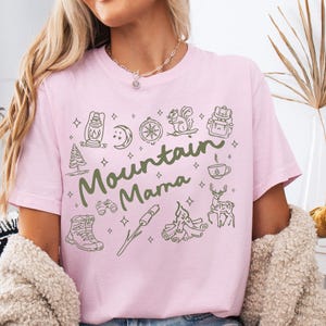May include: A light pink t-shirt with the text "Mountain Mama" in green script, surrounded by outdoor-themed illustrations. The shirt features a crew neck and short sleeves. The word "Blossom" is at the bottom of the image.