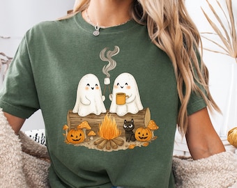 Può includere: T-shirt verde muschio con grafica a tema Halloween. Il design mostra due fantasmi amichevoli seduti vicino a un falò, con zucche, funghi e un gatto nero. La parola "Moss" è stampata in alto a sinistra.