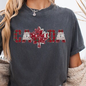 May include: Dark grey t-shirt featuring the word "CANADA" in a patchwork design. The "A" is a red maple leaf with white stars. The letters are red plaid and white lace. The shirt is a Pepper colour.