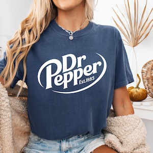 以下が含まれることがあります： 白のDr Pepperロゴが特徴のデニムブルーのTシャツ。ロゴにはブランド名と「Est.1885」が含まれています。このシャツはカジュアルなスタイルで、普段使いに適しています。