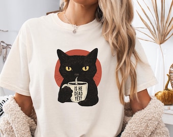 May include: Ivory-colored t-shirt featuring a black cat illustration holding a coffee mug. The cat is set against a red circle, with the text "IS HE DEAD YET?" on the mug. The shirt is a casual wear item.