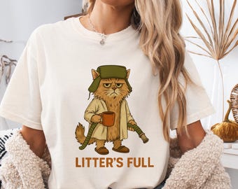 May include: Ivory-colored t-shirt featuring a cartoon cat dressed in a hat and coat, holding a mug and a pipe. The text "LITTER'S FULL" is printed below the cat. The shirt is a relaxed fit.