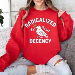 Könnte beinhalten: Ein rotes Sweatshirt mit dem weißen Text "RADICALIZED BY BASIC DECENCY". Eine weiße Taube mit einem Olivenzweig befindet sich unter dem Wort "BY". Das Sweatshirt hat eine einfarbige rote Farbe.