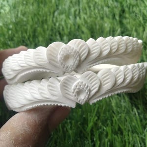 Peut inclure: Deux bracelets blancs finement sculptés. Chaque bracelet présente un motif répété de feuilles stylisées et un motif central en forme de cœur. Les bracelets sont présentés sur un fond d'herbe verte.