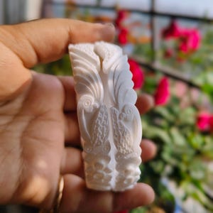 Puede incluir: Un objeto decorativo tallado, de color blanquecino, con intrincados diseños florales y aviformes. El objeto se sostiene en una mano, con un fondo borroso de vegetación y flores. El tallado presenta patrones y texturas detalladas.