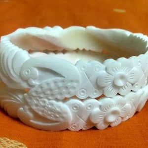 Peut inclure: Deux bracelets blancs sculptés. Les bracelets sont décorés de motifs floraux et d'oiseaux. Le fond est de couleur orange uni. Les bracelets sont empilés l'un sur l'autre.