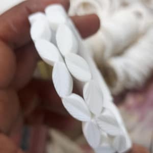 Könnte beinhalten: Ein weißes Armband mit floralem Design. Es besteht aus mehreren weißen, blattförmigen Perlen, jede mit feinen Aderdetails. Die Perlen sind zu einem flexiblen, eleganten Accessoire aufgereiht.