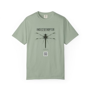 May include: A sage green short-sleeve t-shirt featuring a black graphic of a dragonfly with the text INSECTOTHOPTER, EXPERIMENTAL FLIGHT, and TECHWEAR printed above and below the insect, along with a small square QR code at the bottom centre.