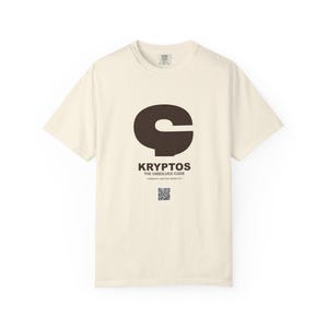 May include: A cream-colored short-sleeve t-shirt featuring a large dark brown graphic of the letter C. Below the graphic, the text reads KRYPTOS THE UNSOLVED CODE, followed by smaller text and a small square QR code centered on the chest area.