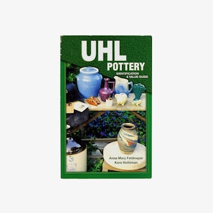 May include: A book titled "UHL Pottery Identification & Value Guide" with a green cover. The cover features various pottery pieces in shades of blue, purple, and white, arranged on a wooden shelf. The book's title is in large white letters.