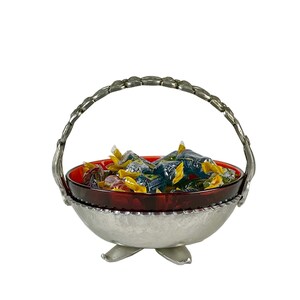 May include: A silver-toned metal basket with a red glass bowl filled with wrapped candies. The basket has a handle and three feet. The candies are in various colors of wrappers.