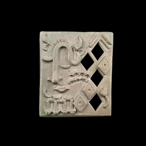 May include: A square, light-green sculpture with a carved face and abstract shapes. The face has a prominent nose, closed eyes, and a curved line for a mouth. Three diamond-shaped cutouts are on the right side. The sculpture has a textured surface.