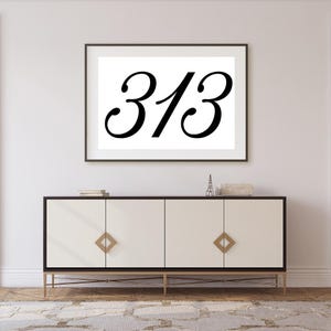 Puede incluir: Una impresión enmarcada con el número "313" en elegante escritura negra. Debajo, un moderno mueble blanco con detalles dorados y tiradores en forma de diamante. El mueble descansa sobre un marco dorado.